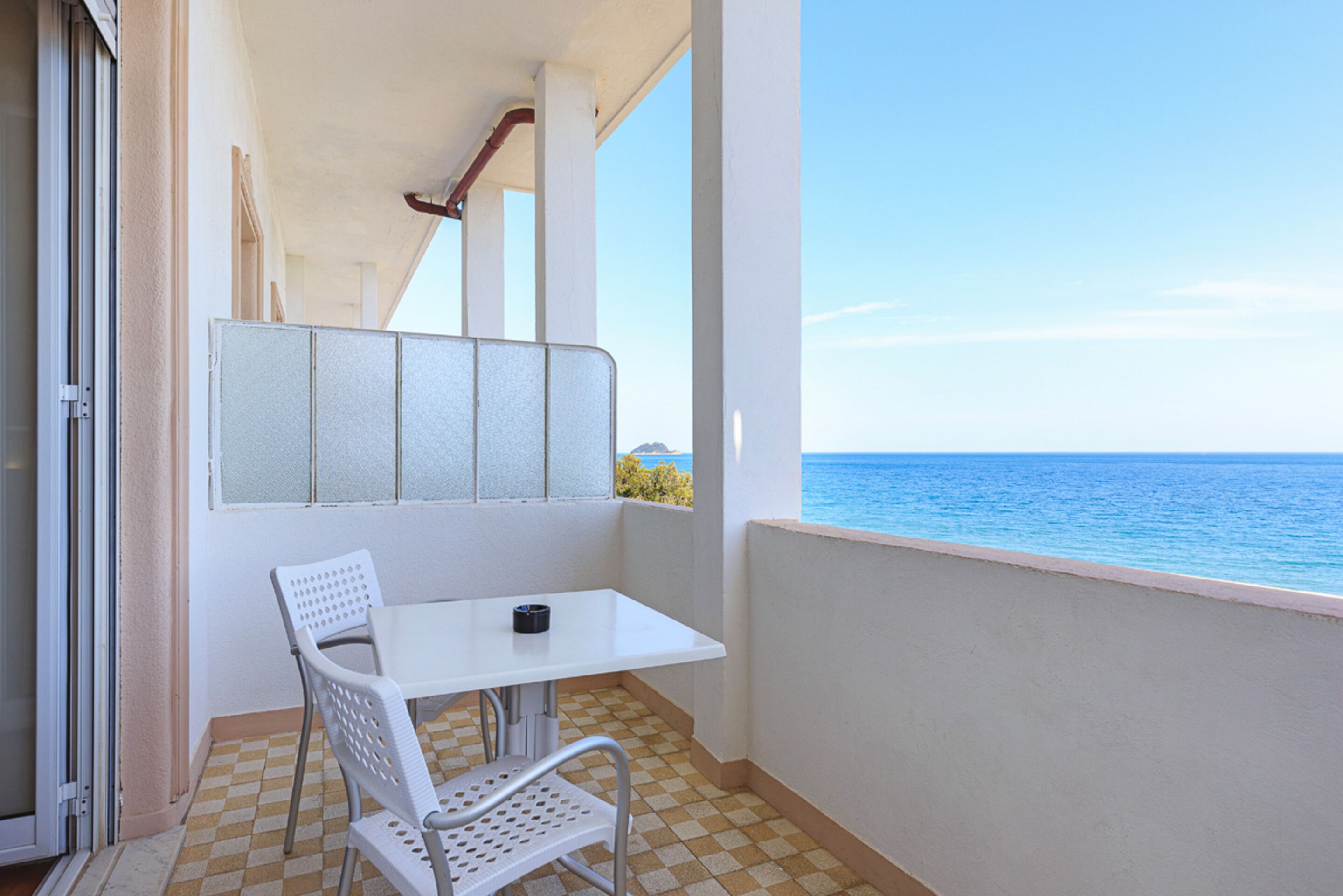 Un balcón con una mesa y silla blancos, vista al mar y piso a cuadros.