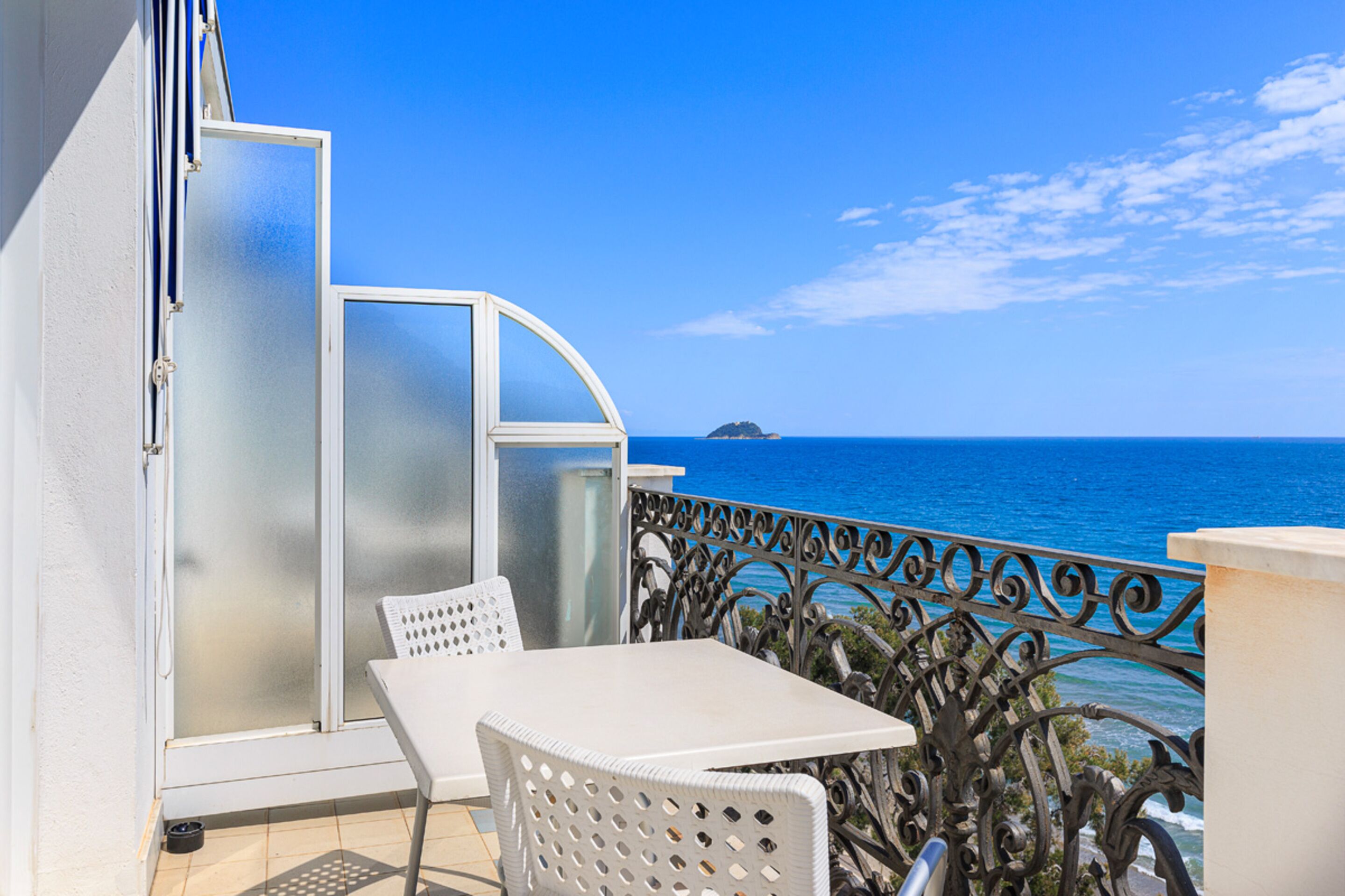 A balcony with a white table and chairs, a glass door, and a view of the sea.