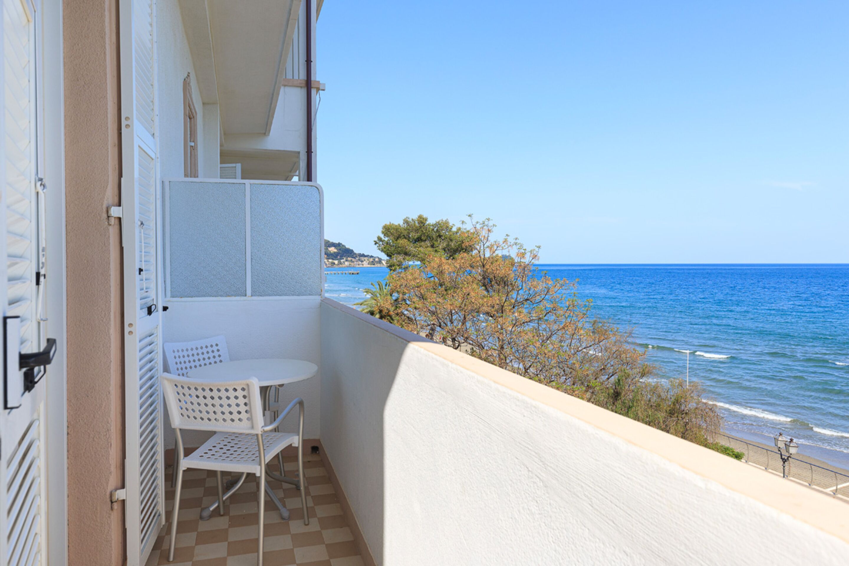 A balcony with a checkered floor, a small table, and two chairs. It overlooks a beach with the sea and a tree in the background.
