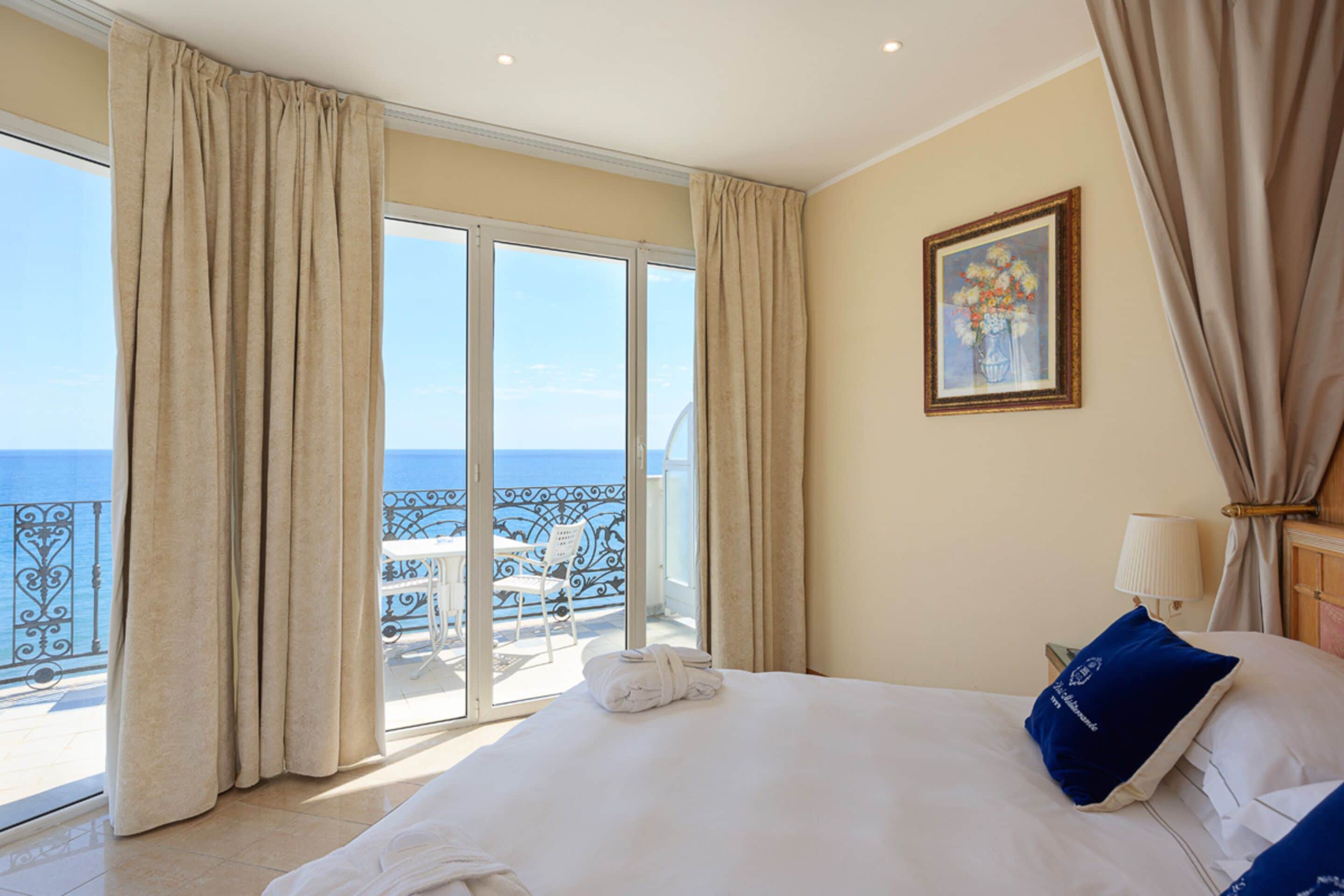 Habitación de hotel con cama, vista al mar, balcón con una silla y un cuadro en la pared.