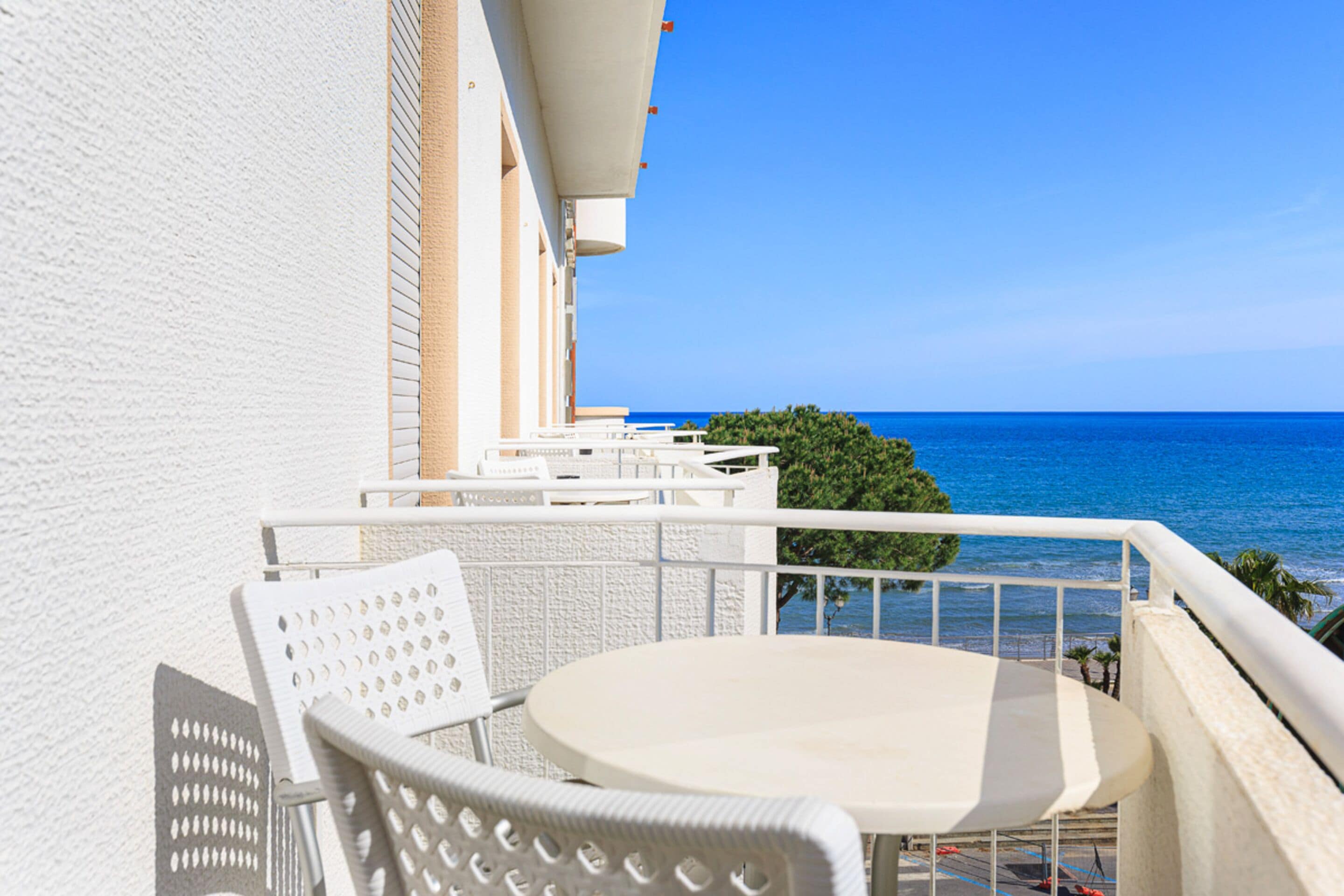 Un balcone con tavolo e sedia bianca, affacciato su un mare azzurro e limpido.