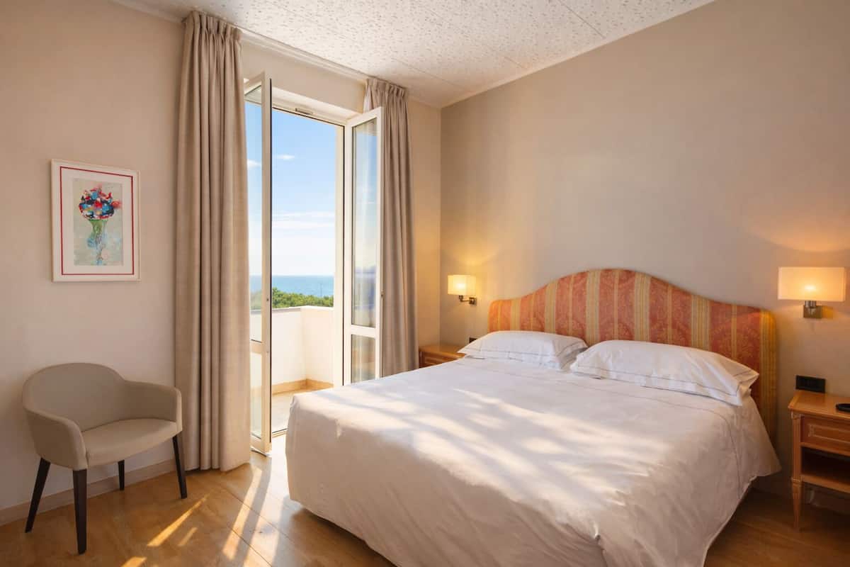 A hotel room with a bed, a chair, a nightstand, a lamp, a painting, and a large window with curtains.