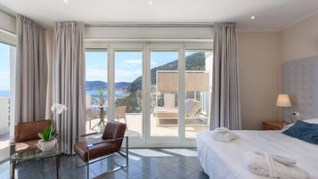 A hotel room with a bed, a seating area with a table and chairs, a potted plant, and a view of the sea through large windows.