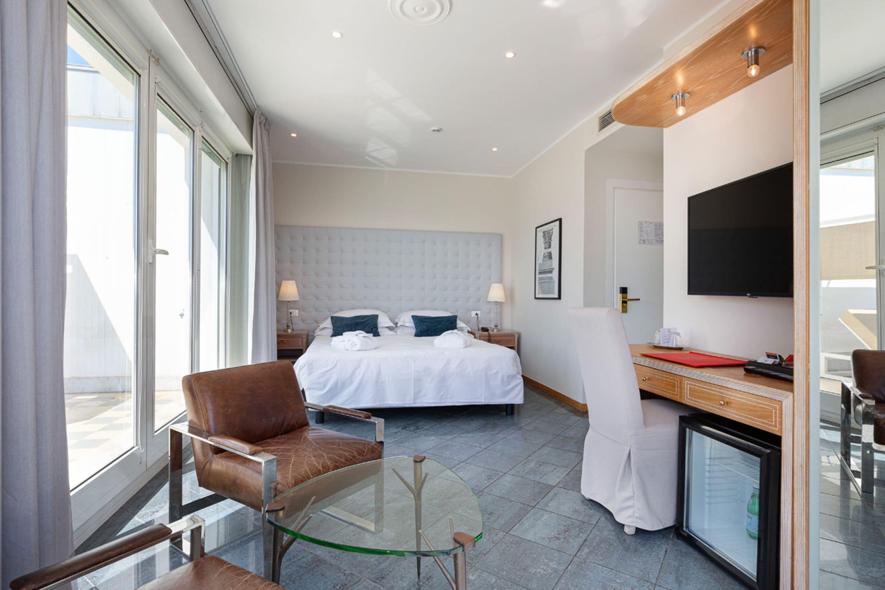 Habitación de hotel con cama, escritorio, silla, mesita de vidrio, televisor y refrigerador pequeño.