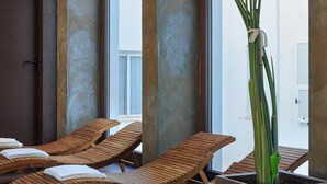 Un área interior luminosa para relajarse, con tumbonas de madera, una planta grande en maceta y vistas amplias a través de grandes ventanales.