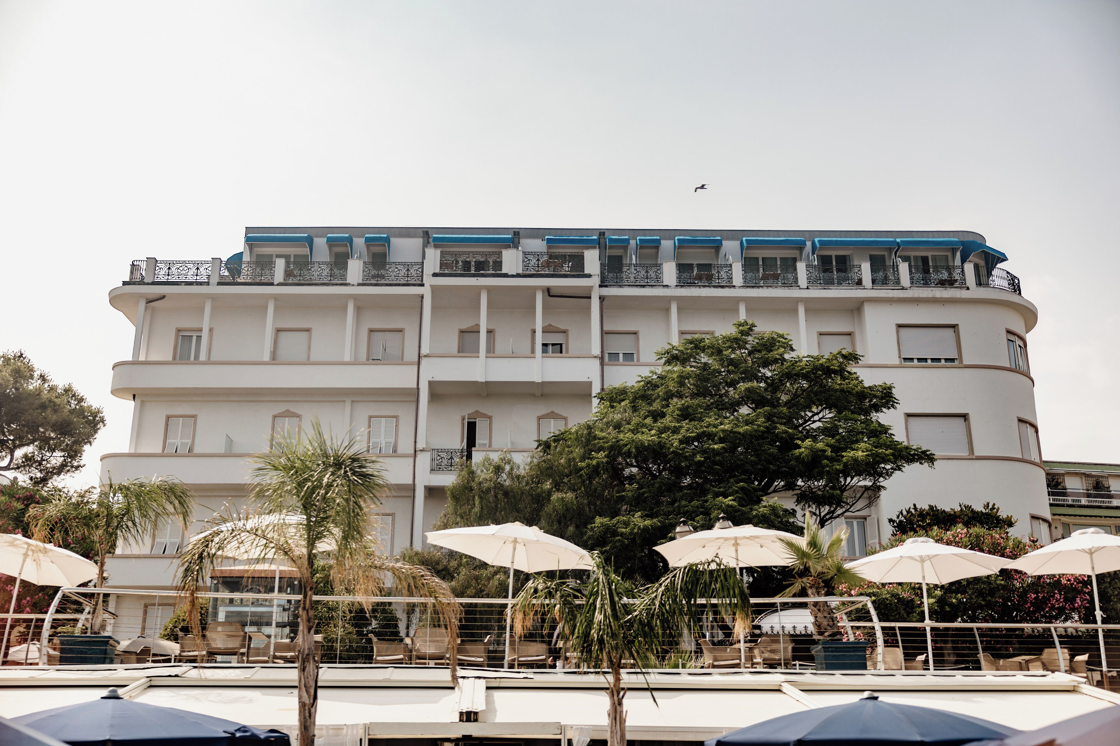 A multi-story hotel with balconies, outdoor seating with umbrellas, and a palm tree.