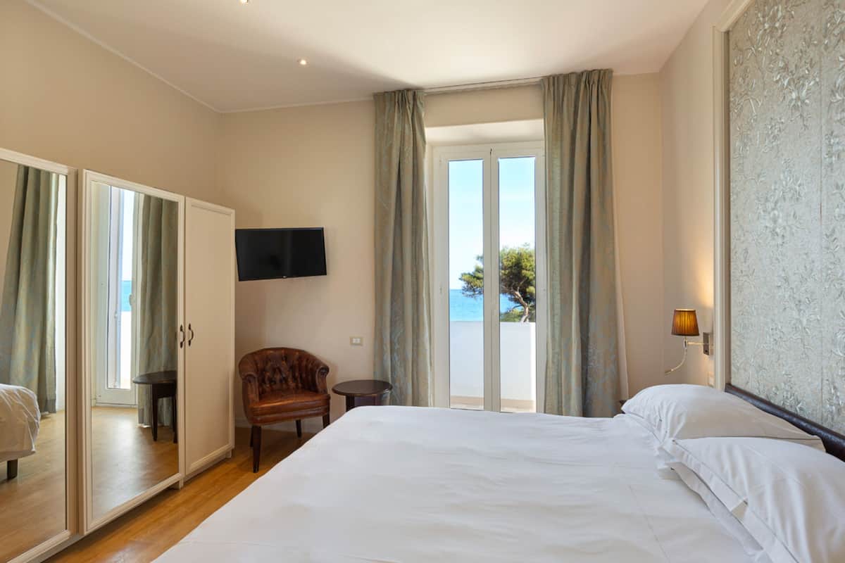 Ein Hotelzimmer mit einem großen Bett, einem Sessel, einem Fernseher und einem Balkon mit Vorhängen.