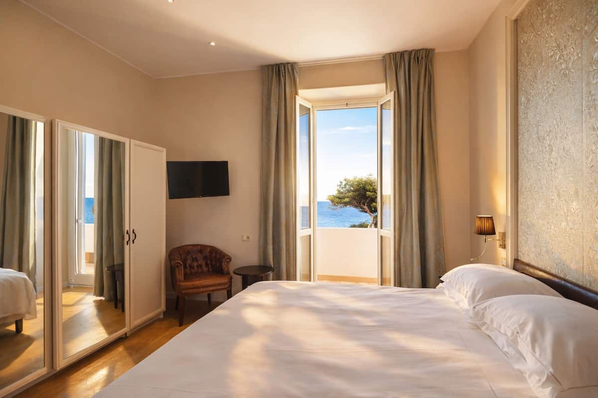 A hotel room with a large bed, a chair, a TV, and a balcony with a view of the sea.