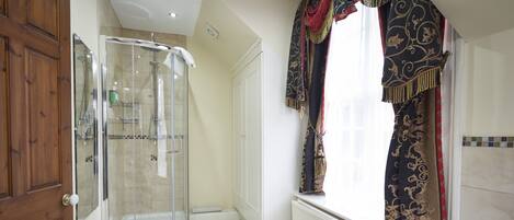 A bathroom with a glass shower enclosure, a wooden door, and a window with ornate curtains.