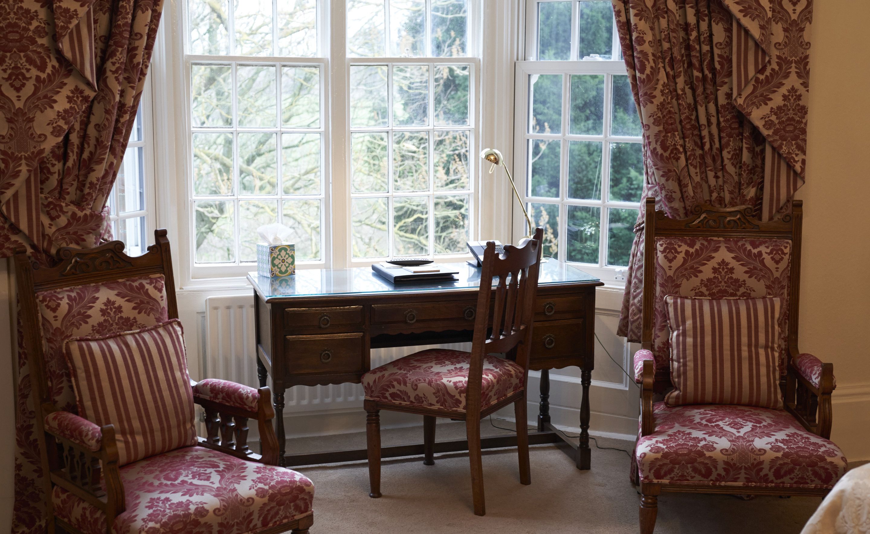 A room with a wooden desk, two chairs, a radiator, and a window with curtains.