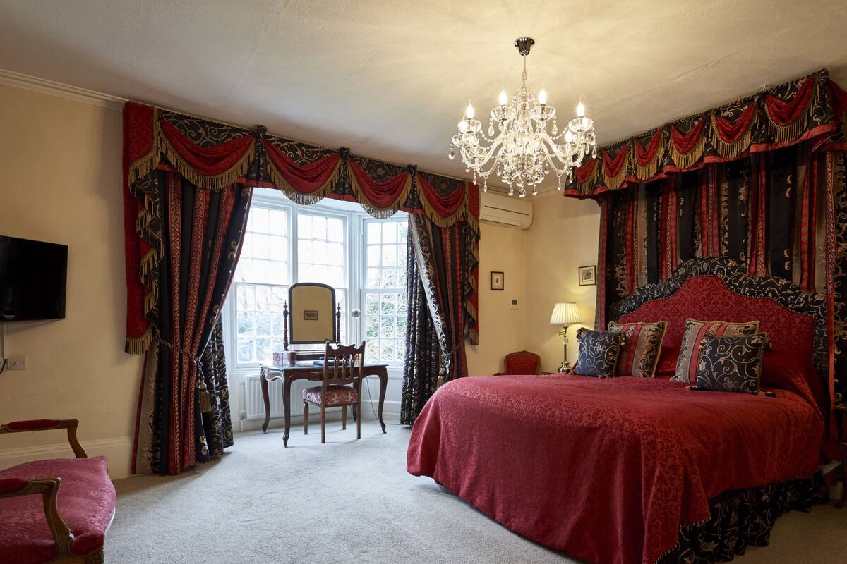 A bedroom with a large bed, a chandelier, and heavy drapery.