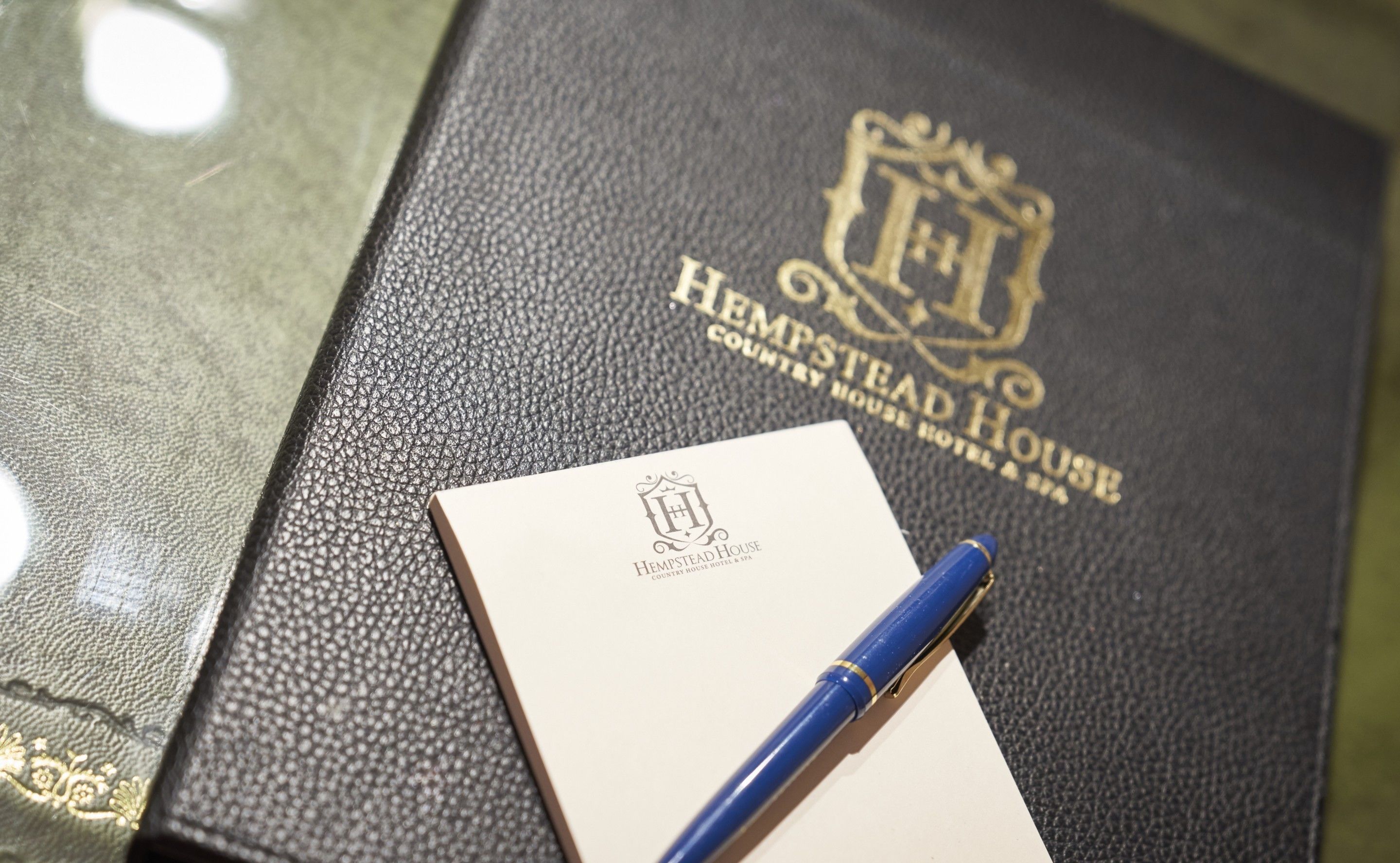 A black leather-bound book with gold embossed text reading "HEMPSTEAD HOUSE COUNTRY HOUSE HOTEL & SPA". A blue pen rests on a white card with the same embossed logo.