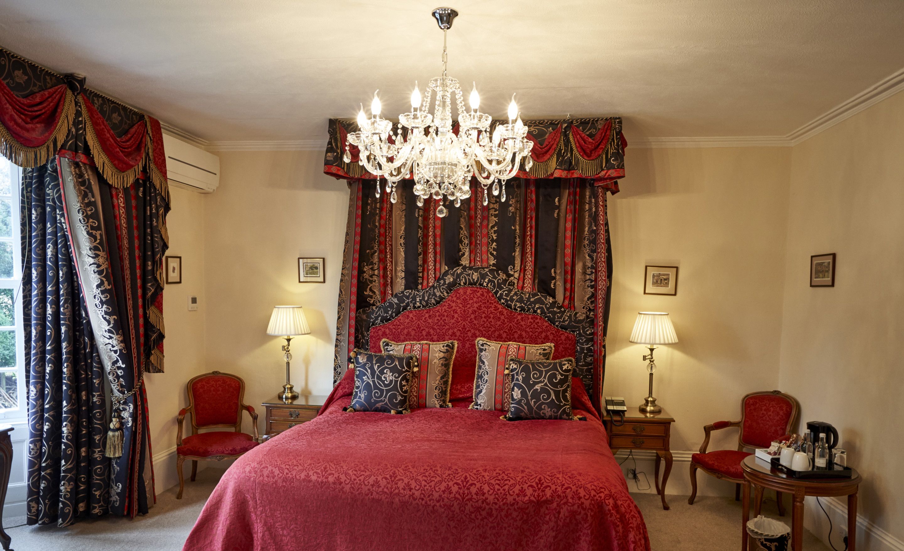 A spacious bedroom with a large bed, ornate curtains, a chandelier, and classic furniture.