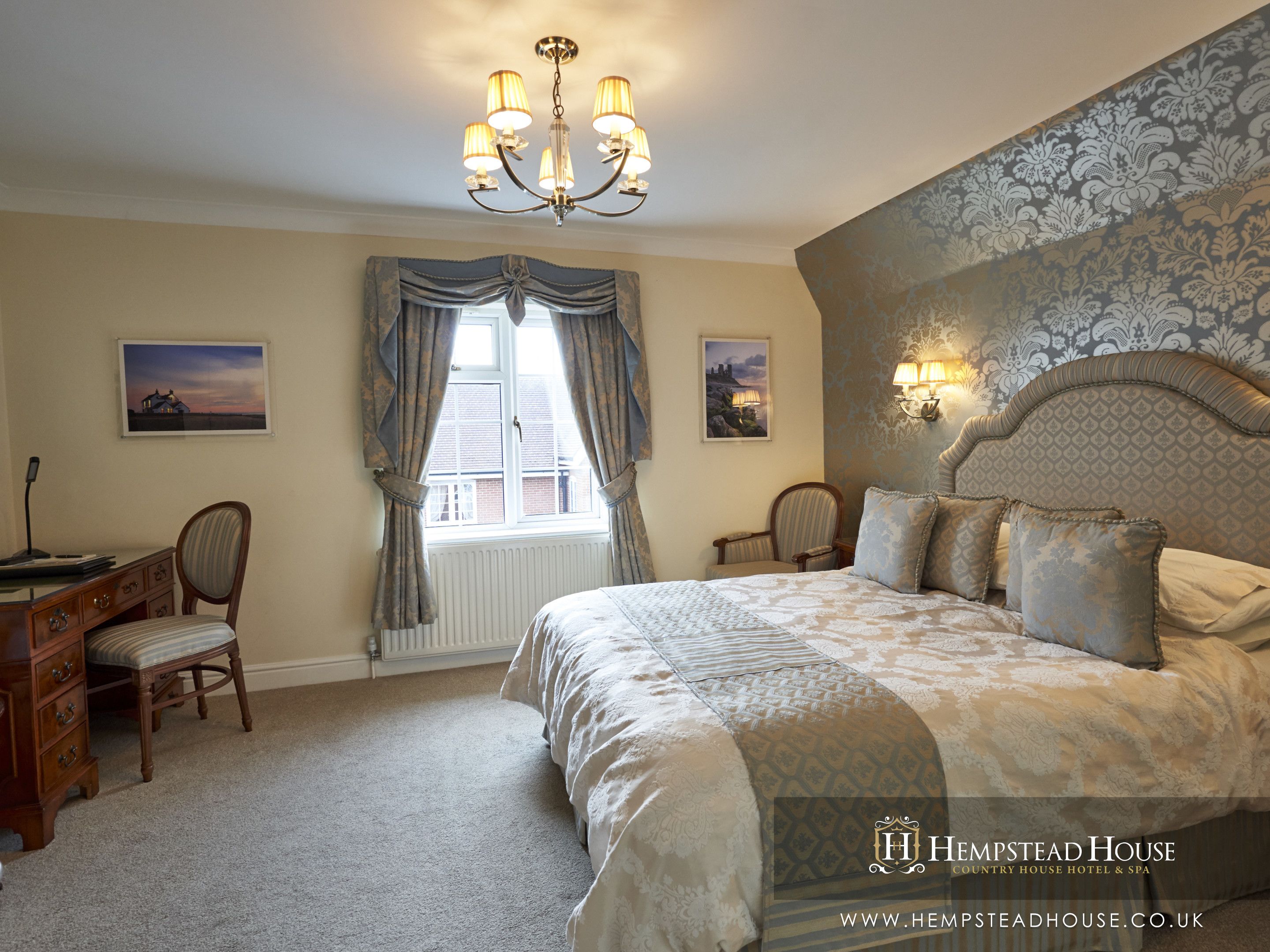 A bedroom with a large bed, a desk, and two chairs. There are two windows with curtains, a chandelier, and wall-mounted lamps.