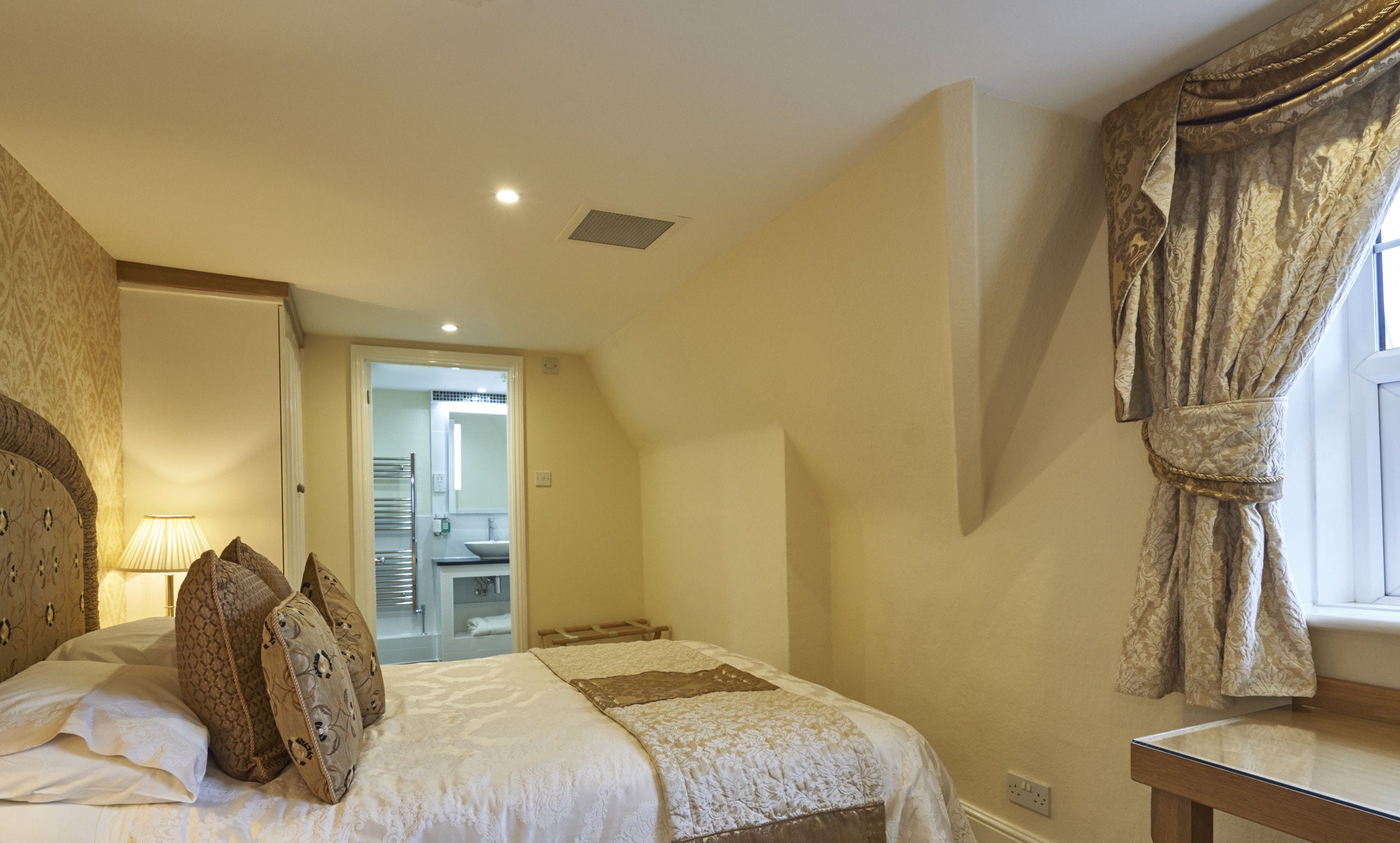 A bedroom with a bed, a nightstand, a lamp, a window with curtains, and a bathroom visible through an open door.