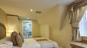 A bedroom with a bed, a nightstand, a lamp, a window with curtains, and a bathroom visible through an open door.