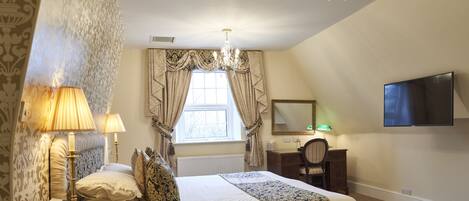 A bedroom with a large bed, two bedside lamps, a chandelier, a window with curtains, a desk with a chair, a mirror, and a TV mounted on the wall.