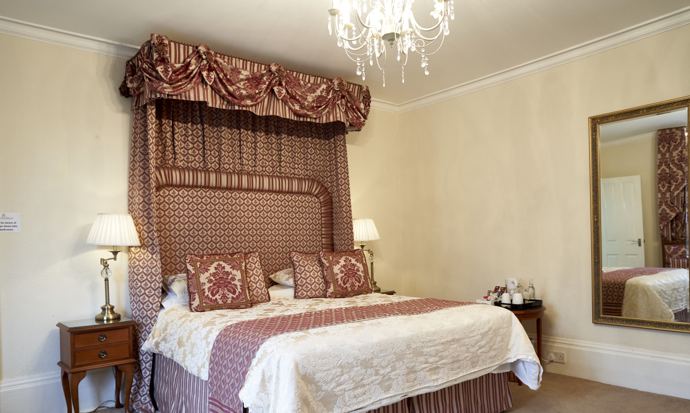 A four-poster bed with a patterned canopy, two bedside lamps, a mirror, and a nightstand with a lamp.
