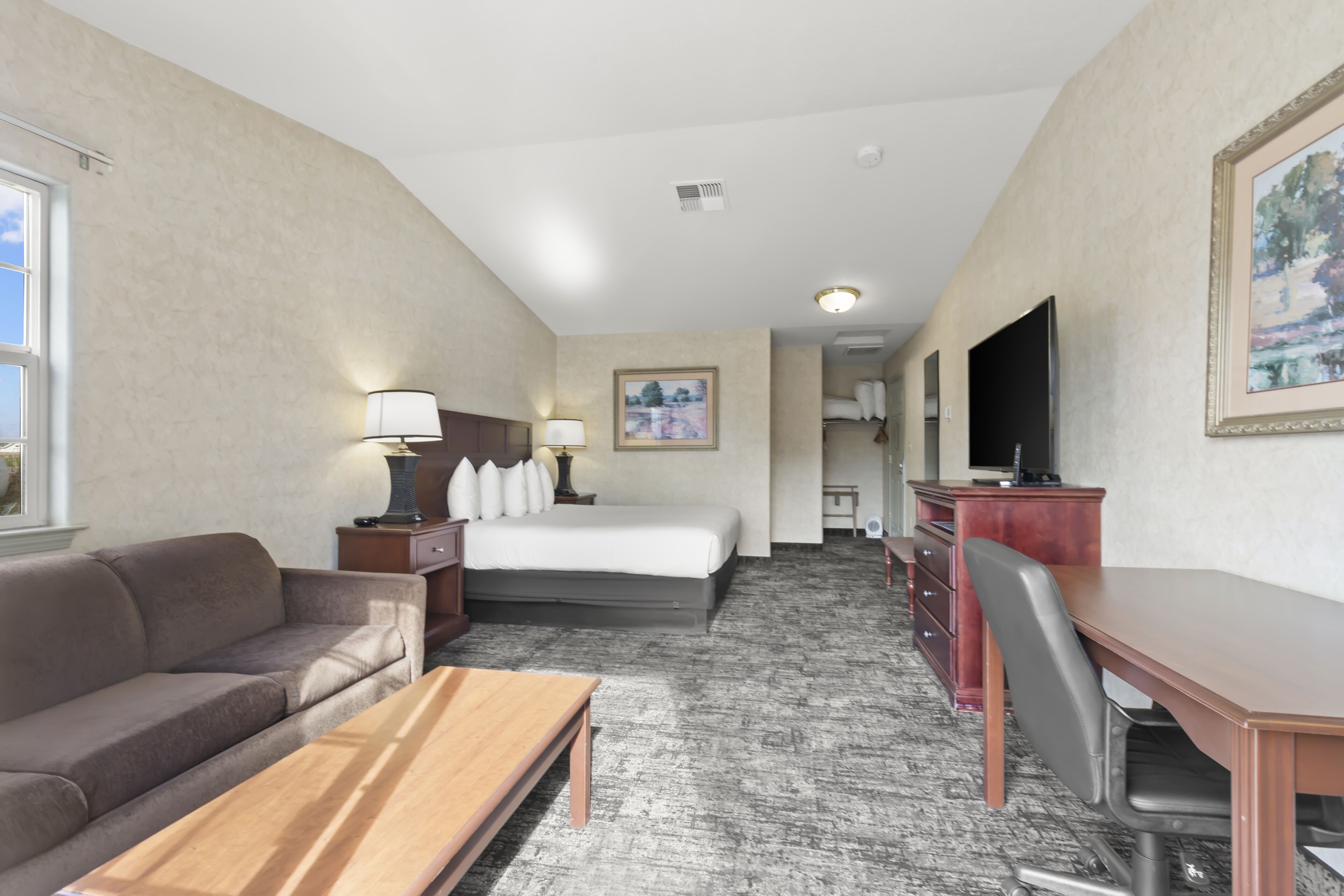 A hotel room with a bed, a desk, a chair, a sofa, a TV, and a painting on the wall.