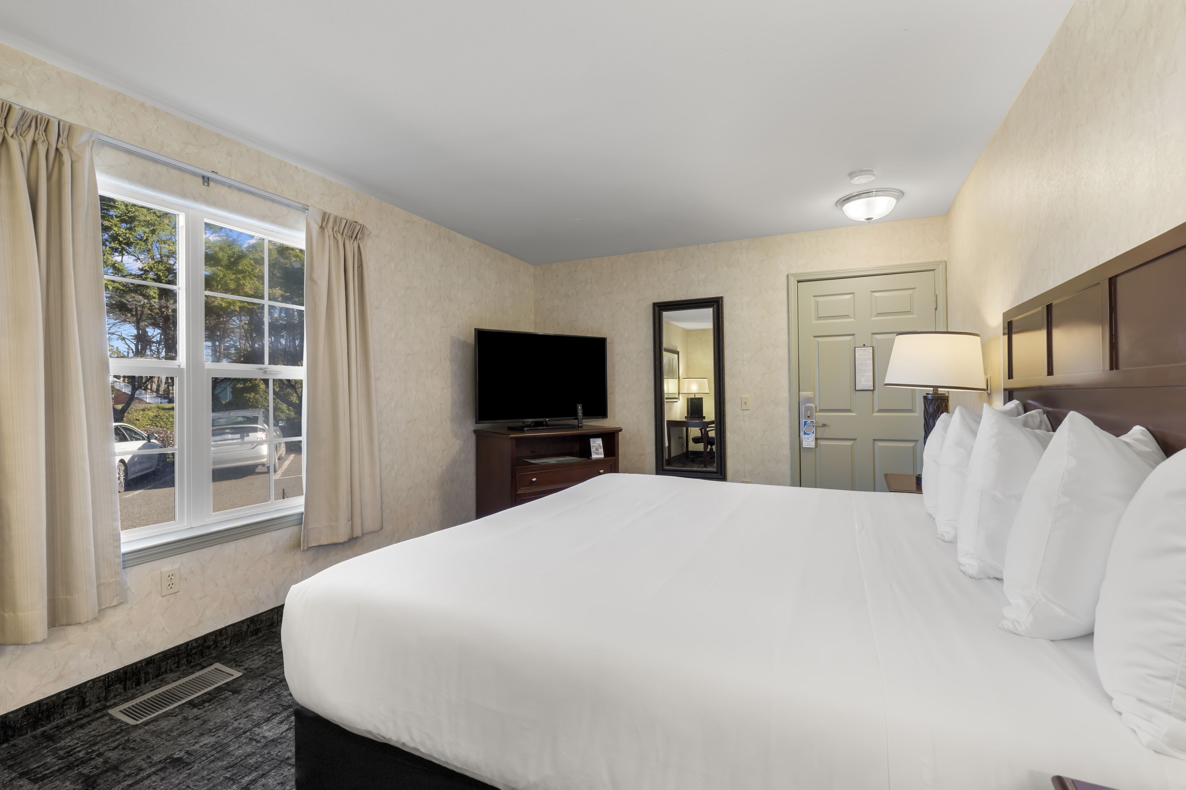 A hotel room with a large bed, a flat-screen TV, a desk, a lamp, and a window with curtains.