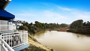 A riverside view with a wooden deck and railing, surrounded by greenery and a clear sky.
