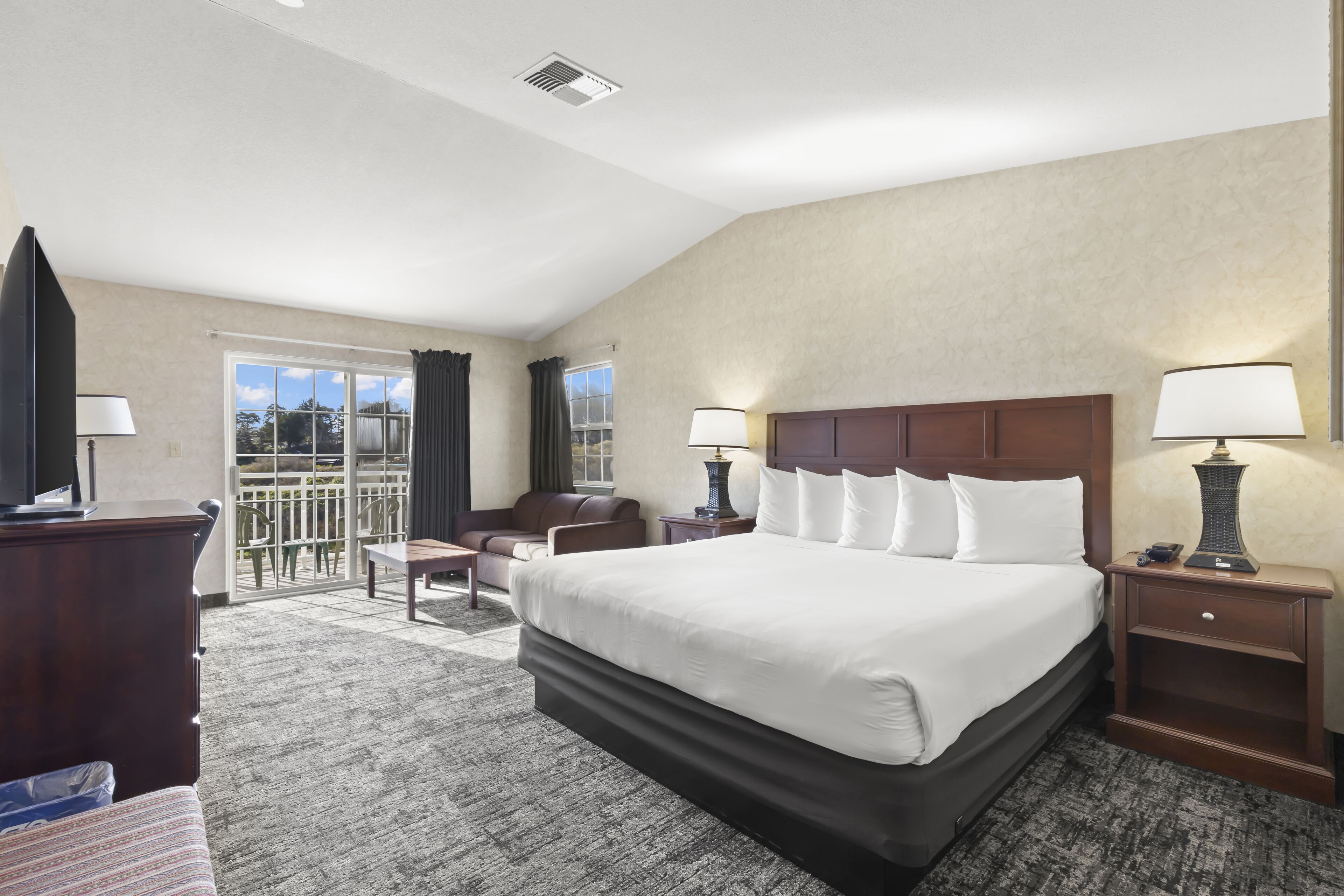 A hotel room with a large bed, a desk, a chair, a TV, and a balcony with a view.