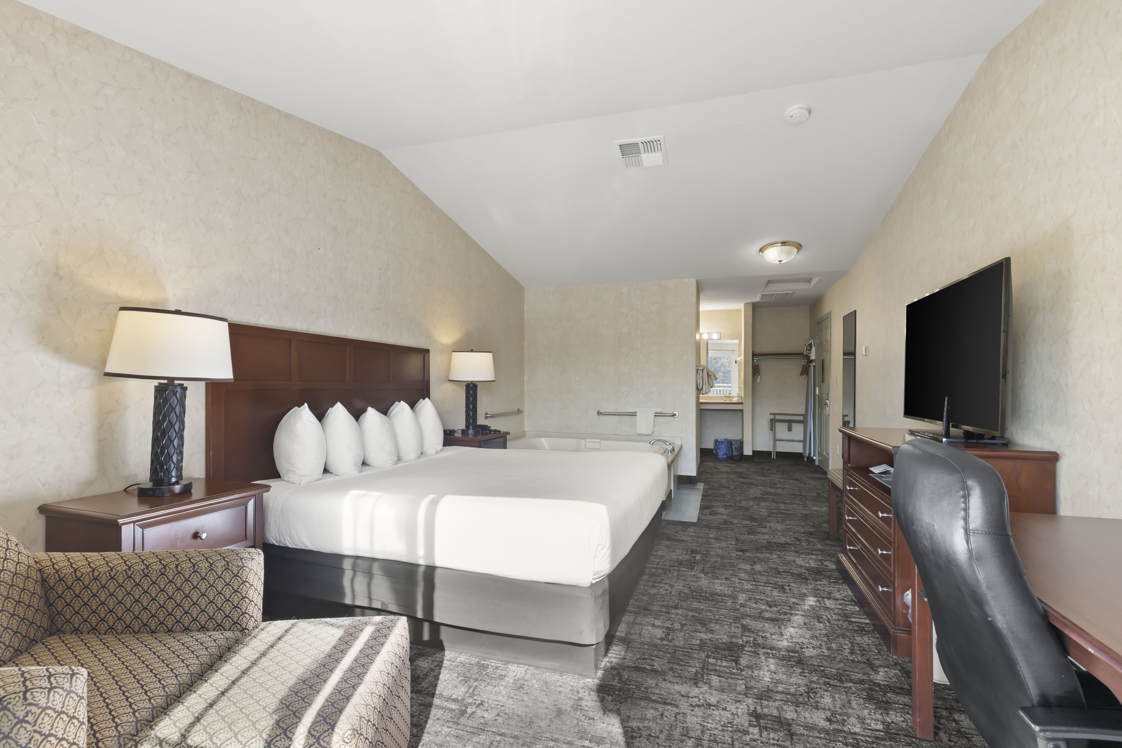 A hotel room with a large bed, a desk with a chair, a television, and a bathroom visible in the background.