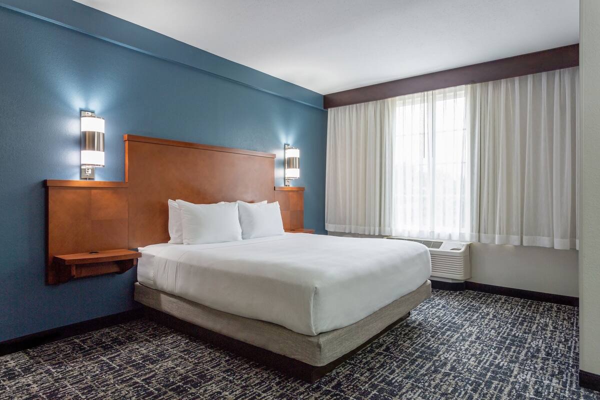 A hotel room with a large bed, two bedside tables, a nightstand, a wall-mounted lamp, and a window with curtains.
