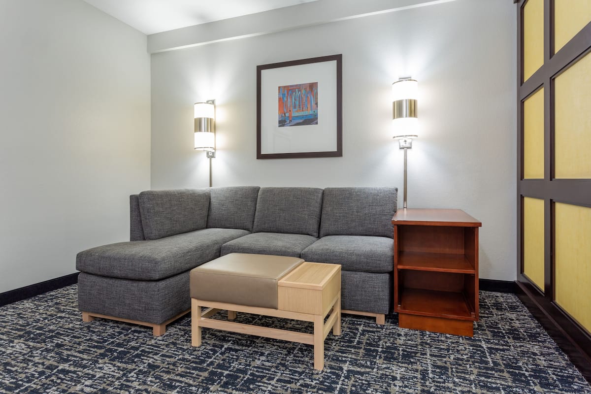 A modern living room with a grey sectional sofa, a wooden coffee table, and a small wooden side table. There is a framed artwork on the wall and two wall-mounted lamps.