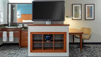 Habitación de hotel con un televisor de pantalla plana sobre un soporte de madera, un mueble de madera con un objeto azul, una mesa de madera con una silla y un baño visible a través de una puerta abierta.