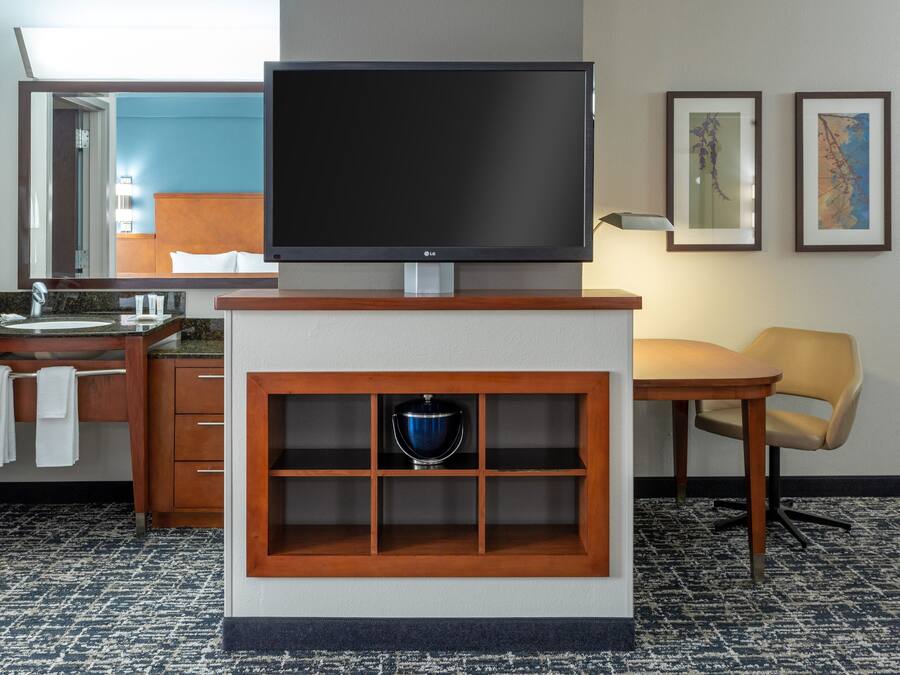 Habitación de hotel con un televisor de pantalla plana sobre un soporte de madera, un mueble de madera con un objeto azul, una mesa de madera con una silla y un baño visible a través de una puerta abierta.