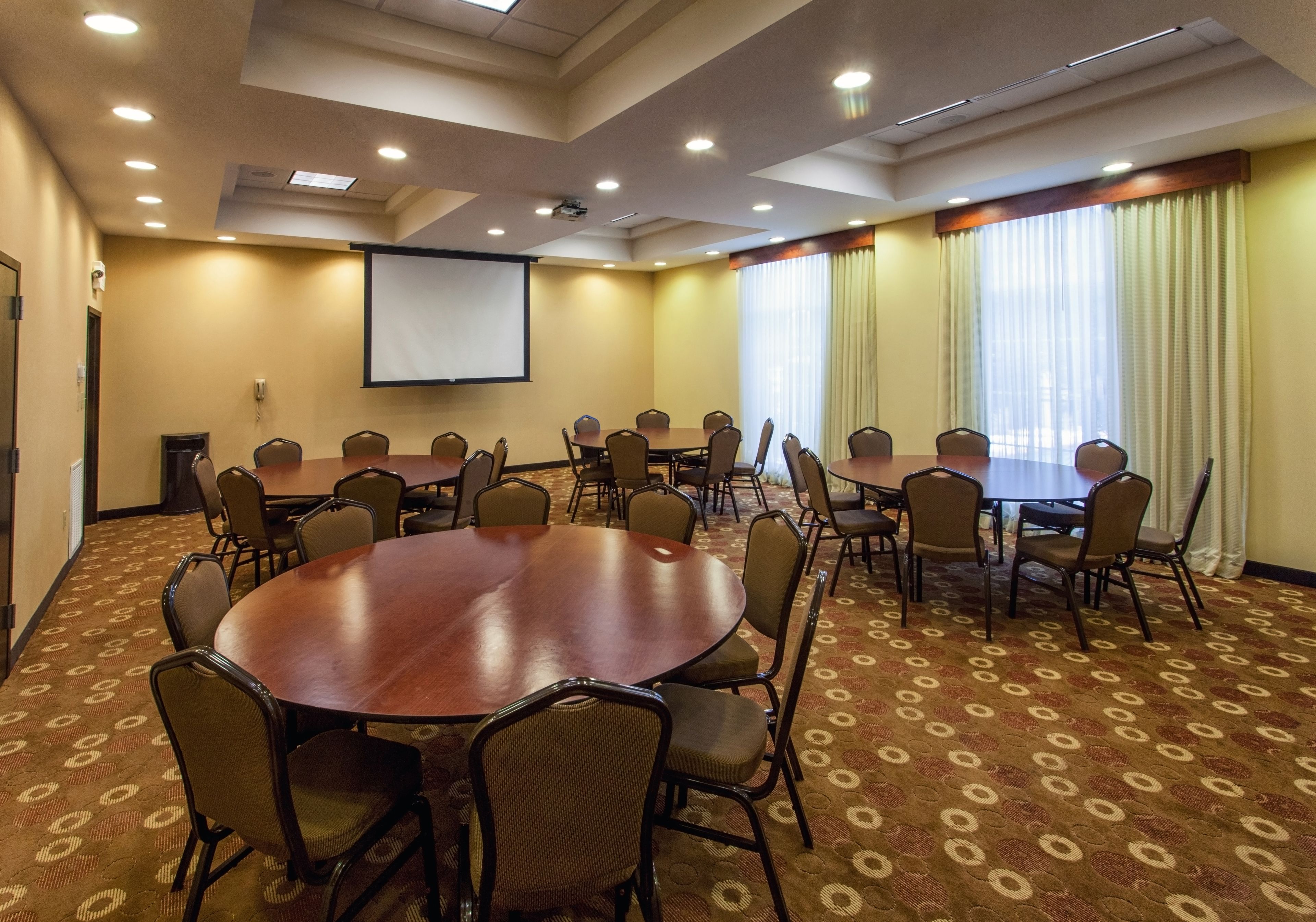 A conference room with a large oval table surrounded by chairs, a projector screen, and a carpeted floor.
