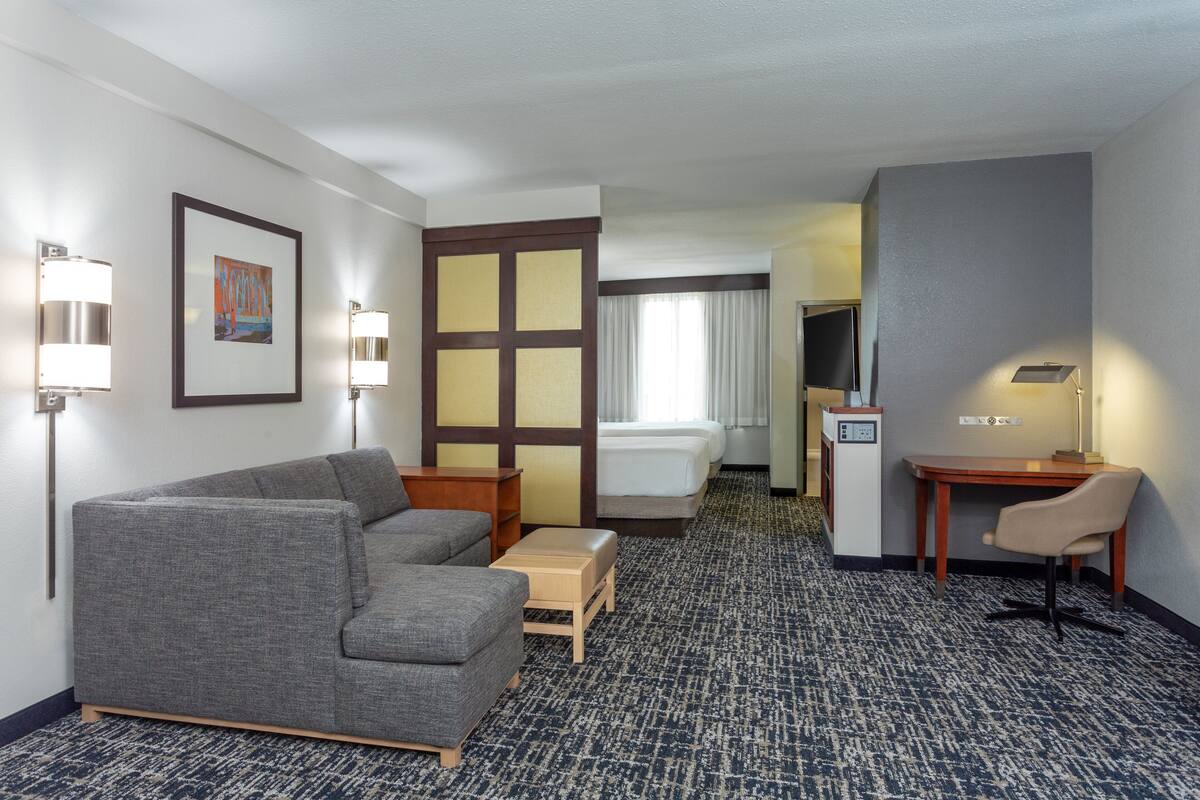 A hotel room with a gray sofa, a small table, a desk with a chair, a bed, and a TV.