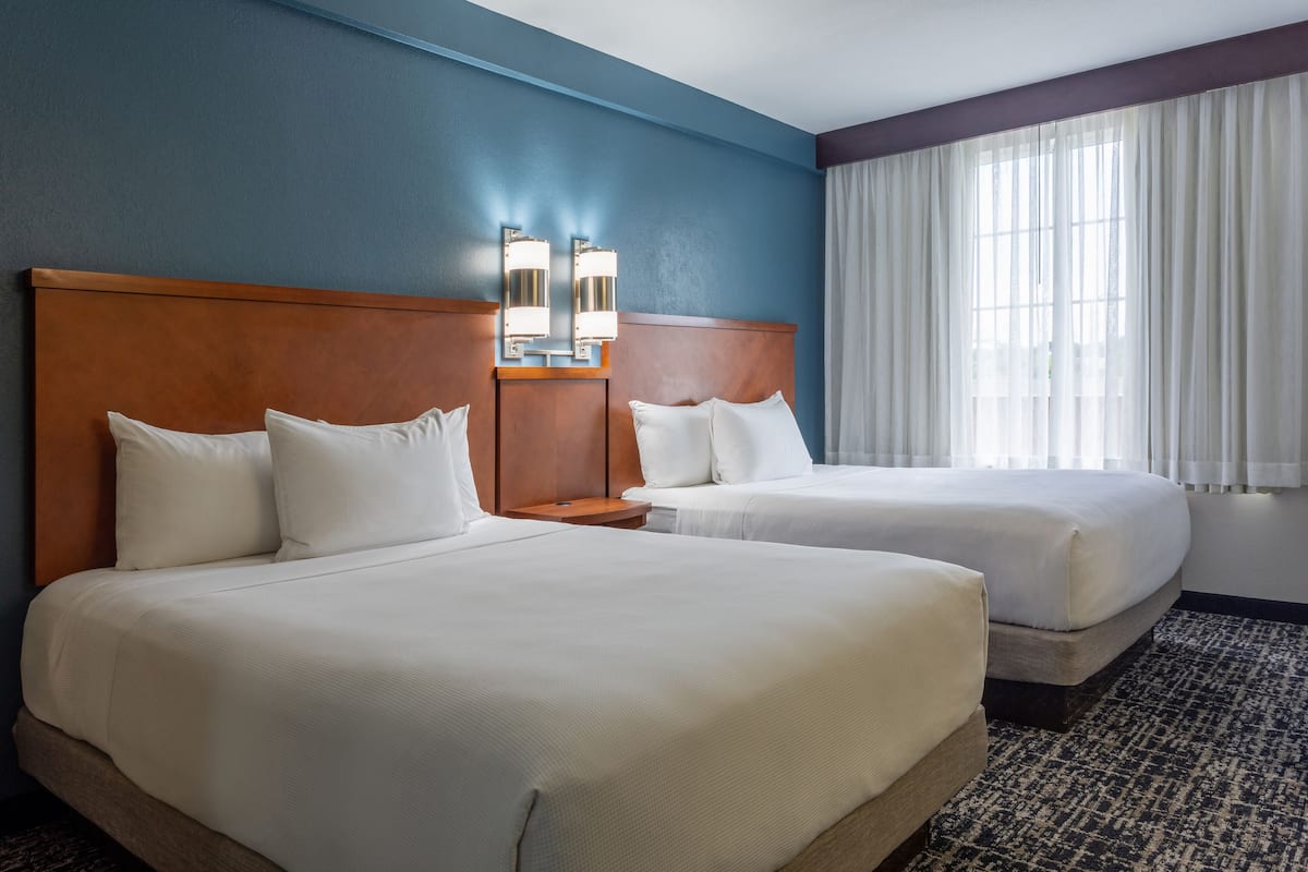 A hotel room with two beds, a wooden headboard, a nightstand, and a large window with curtains.