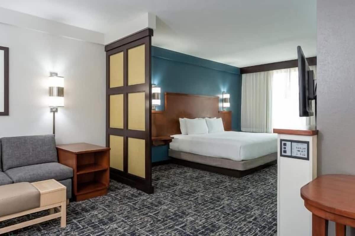 A hotel room with a bed, a sofa, a desk, a TV, and a wall-mounted control panel.