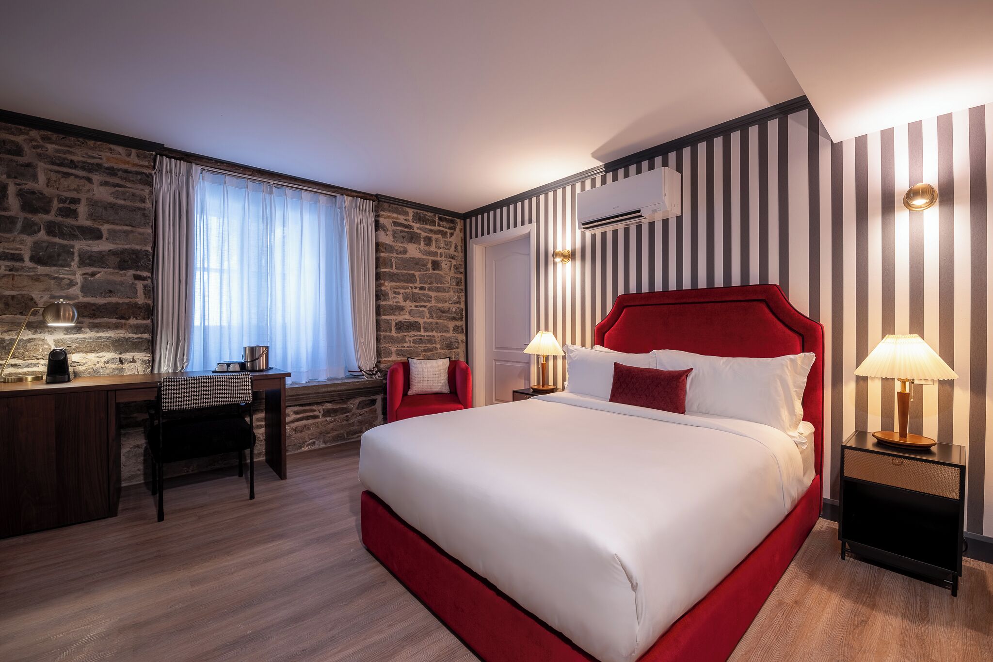 A hotel room with a large bed, a desk, a chair, and a stone wall.