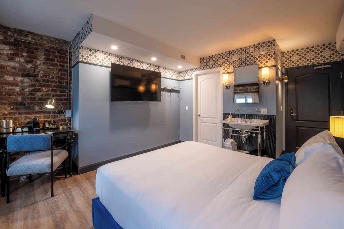 A hotel room with a bed, a desk with a chair, a TV, a sink, and a door.
