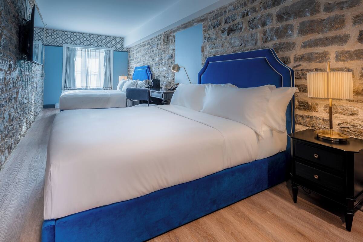 A hotel room with a large bed, a desk with a chair, a lamp, and a stone wall.