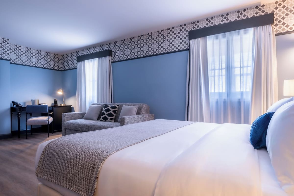 A hotel room with a large bed, a desk with a chair, and two windows with sheer curtains.