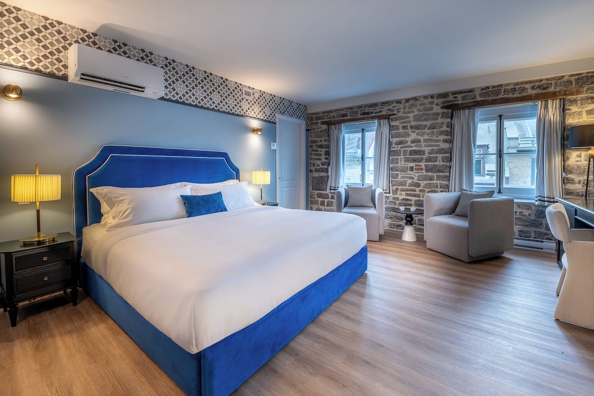 A modern hotel room with a large bed, a stone accent wall, and a view of the city through the window.