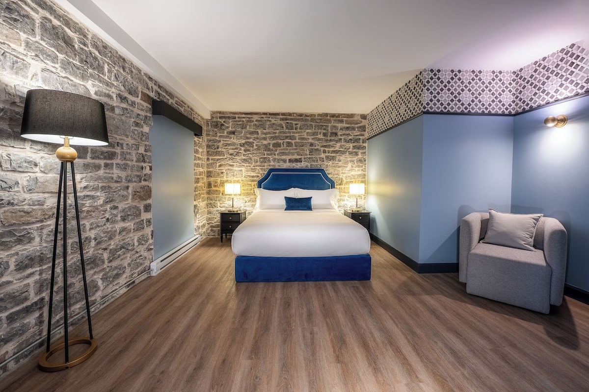 A bedroom with a stone wall, a bed with a blue headboard, a side table with a lamp, and a grey armchair.