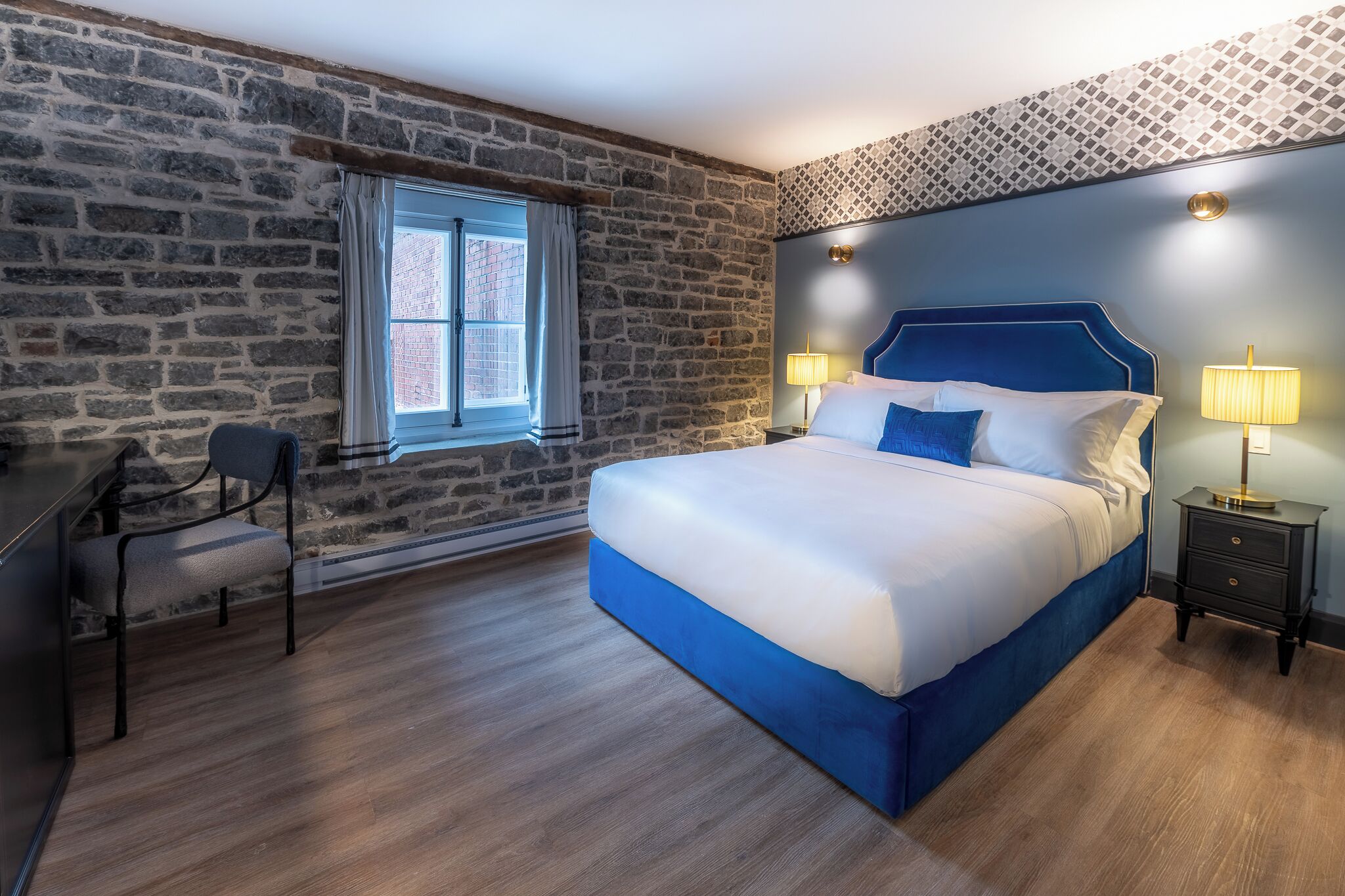 Un dormitorio con pared de piedra, cama con cabecera azul, escritorio con silla y ventana con cortinas.