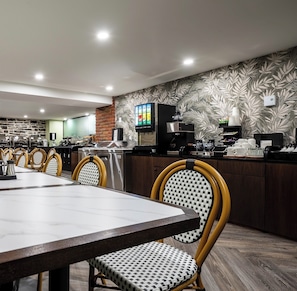 A modern dining area with a long table, wicker chairs, a brick accent wall, and a kitchen area with appliances.