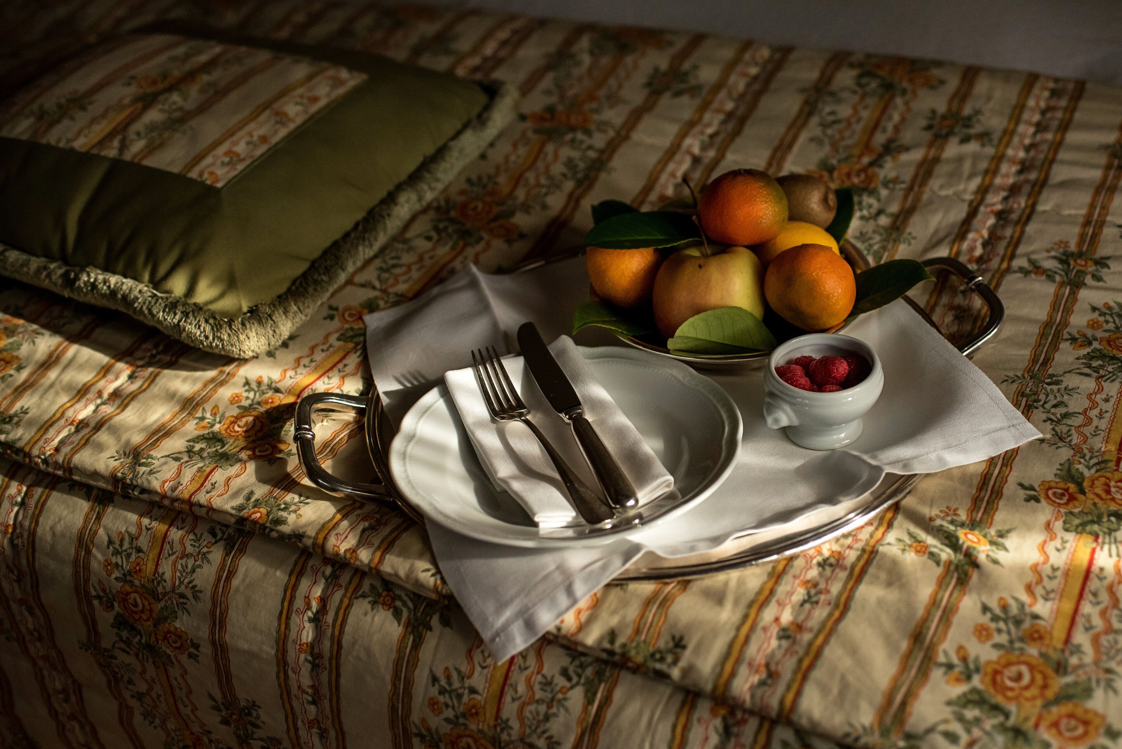 A neatly arranged breakfast setting on a bed with a floral-patterned bedspread, a tray with fruits, and cutlery.
