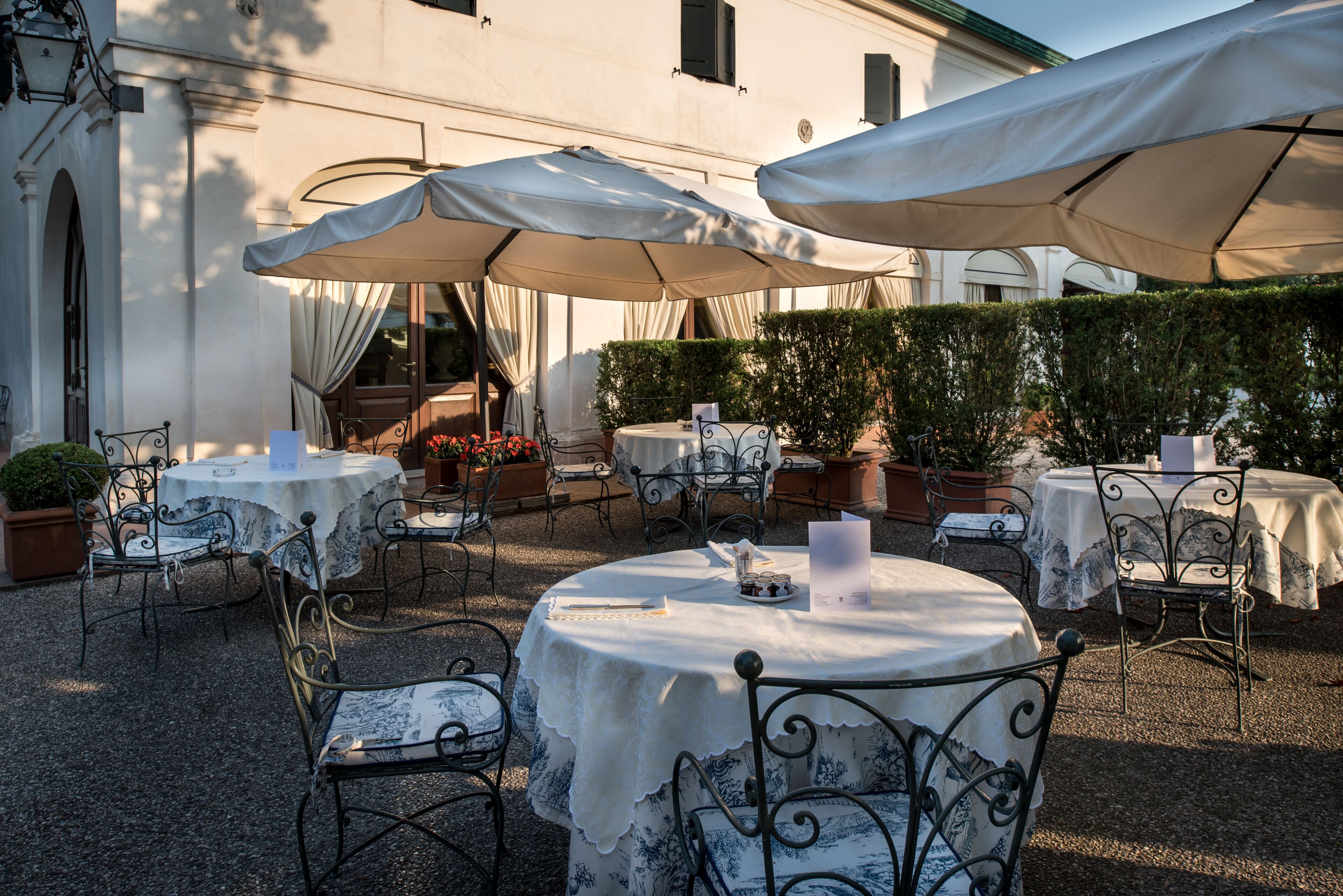 An outdoor patio with round tables and chairs, covered by large umbrellas, and surrounded by potted plants and a building facade.