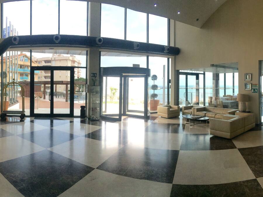 A hotel lobby with a checkered floor, a reception desk, a seating area with sofas, and large windows offering a view of the cityscape.