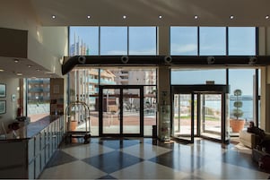 Un vestíbulo de hotel con mostrador de recepción, puerta de cristal de entrada y vistas a la ciudad y al agua.