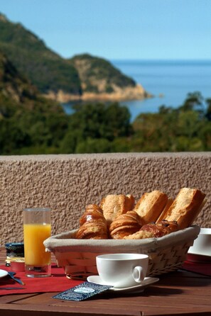 A breakfast setting with bread, pastries, and a glass of orange juice on a table with a scenic view of the sea and hills. - Hôtel Capo D'Orto (Ota)