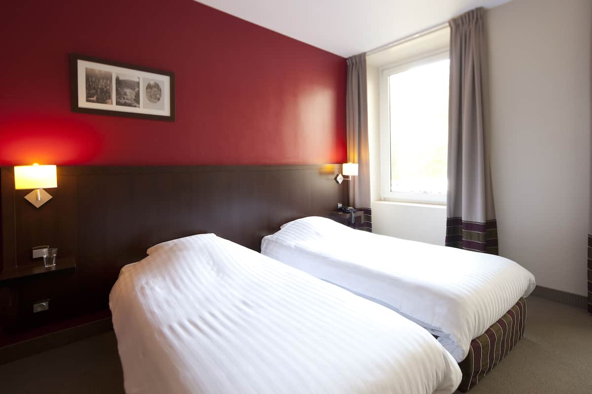 Een hotelkamer met twee bedden, een rode wand, een lijst met een afbeelding en een raam met gordijnen.
