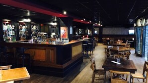 Un espace bar avec un comptoir, des tabourets et des tables. On trouve des étagères remplies de bouteilles et de verres derrière le comptoir.